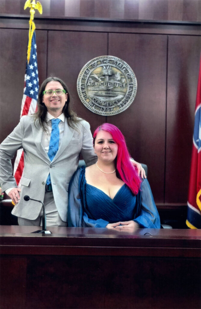 A photo of Lauryn Hills sitting in the judge’s chair in front of the Great Seal of Tennessee. Joe Hills leans jauntily on the chair. 