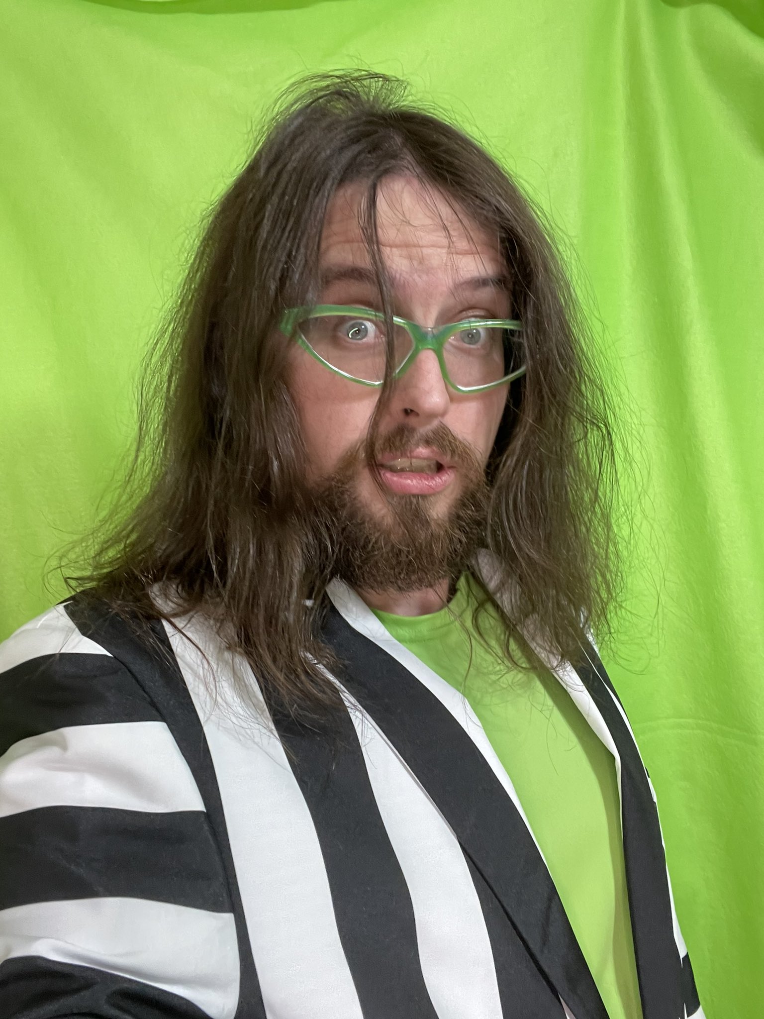 A photograph of Joe Hills with unkempt hair wearing a black and white striped suit jacket over a green tank top 